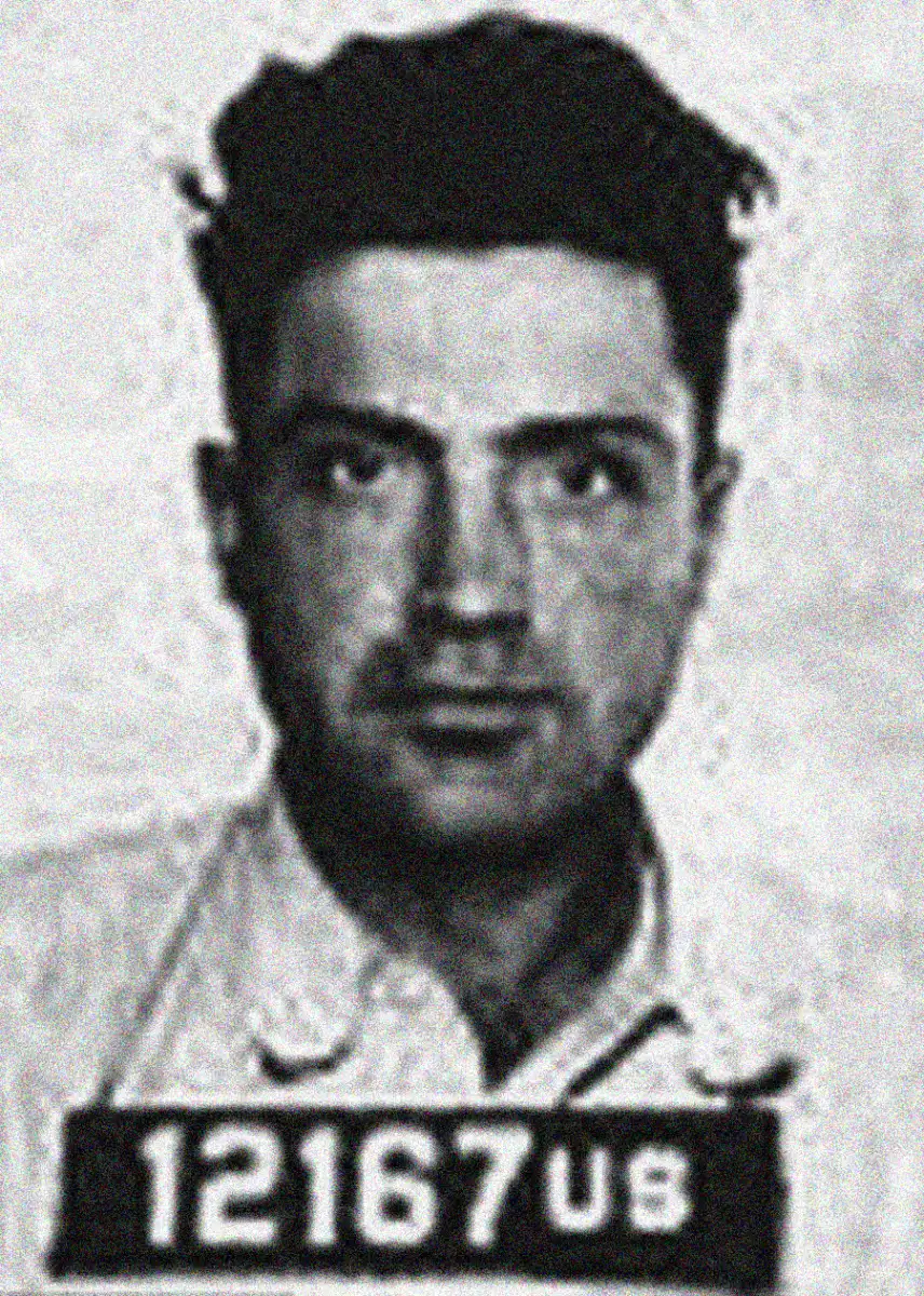 Black and white mugshot of a man with dark hair, wearing a light-colored shirt. He is looking directly at the camera, and there is a sign with the number "12167 UB" visible below his face.
