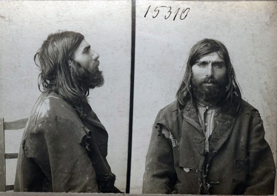 Black and white mugshot of a bearded man with long hair, wearing a tattered coat. The photo shows both a side profile (left) and a front view (right), with the number 15310 written at the top.