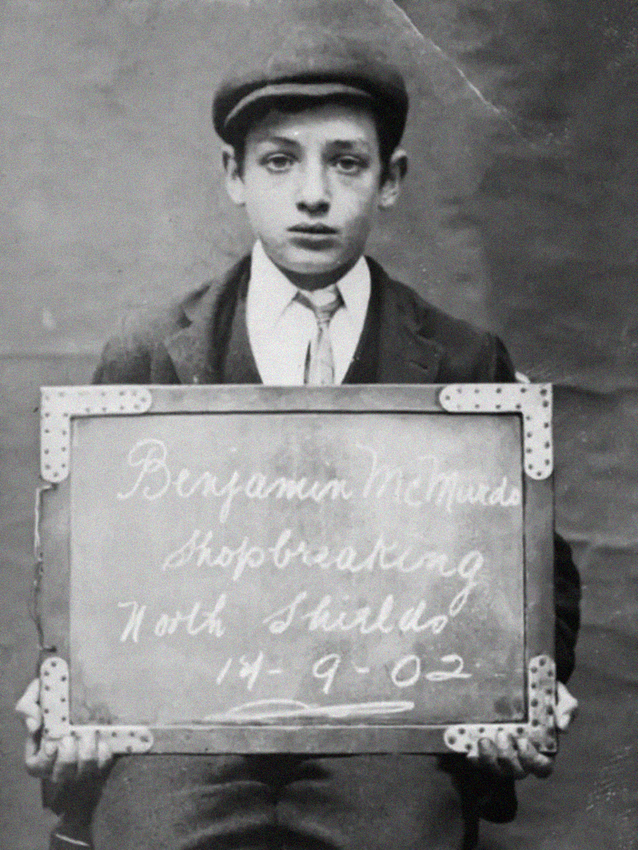A young boy wearing a flat cap and suit holds a chalkboard sign reading "Benjamin McMards, shopbreaking, North Shields, 14-9-02" in a black and white vintage photograph.