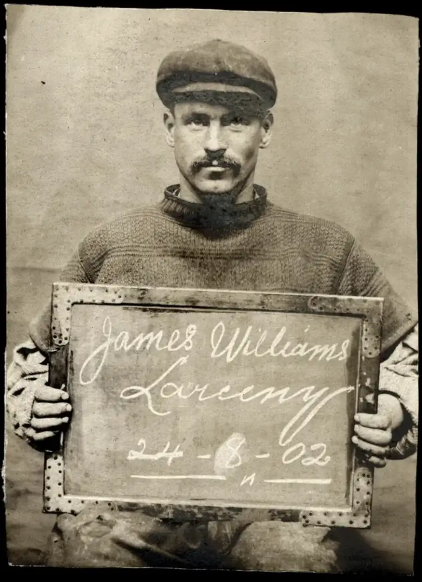 Black-and-white photo of a man in a cap and sweater holding a sign that reads “James Williams, Larceny, 24-8-02.” The background is plain and the image has an old, vintage appearance.