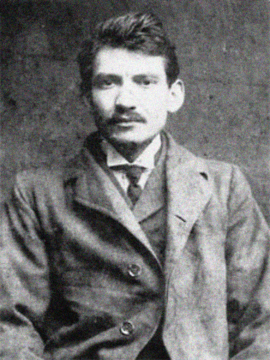 Black and white photo of a young man with short, dark hair and a mustache, wearing a suit and tie, sitting in front of a plain backdrop and looking directly at the camera.