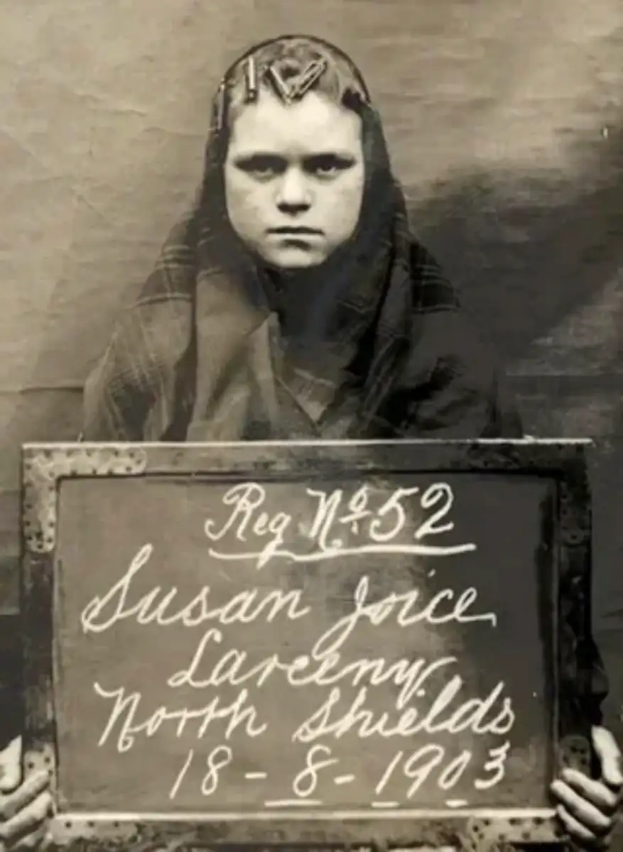 Black-and-white photograph of a young girl wearing a plaid shawl, holding a sign that reads "Reg No 52, Susan Joyce, Larceny, North Shields, 18-8-1903." She stares seriously at the camera.