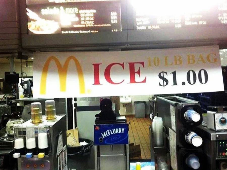 A large sign inside a fast food restaurant advertises "ICE 10 LB BAG $1.00" with the McDonald’s golden arches logo forming the "M" in "ICE." Restaurant equipment and a McFlurry machine are visible below the sign.