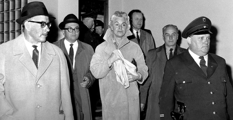 A black-and-white photo shows a woman in a light coat walking between men in suits, hats, and an officer in uniform. The group appears to be indoors, surrounded by other people in the background.