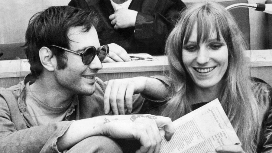A man wearing sunglasses sits next to a smiling woman holding a newspaper. They appear relaxed and happy, seated closely together. A person in the background is partially visible, dressed in formal attire.