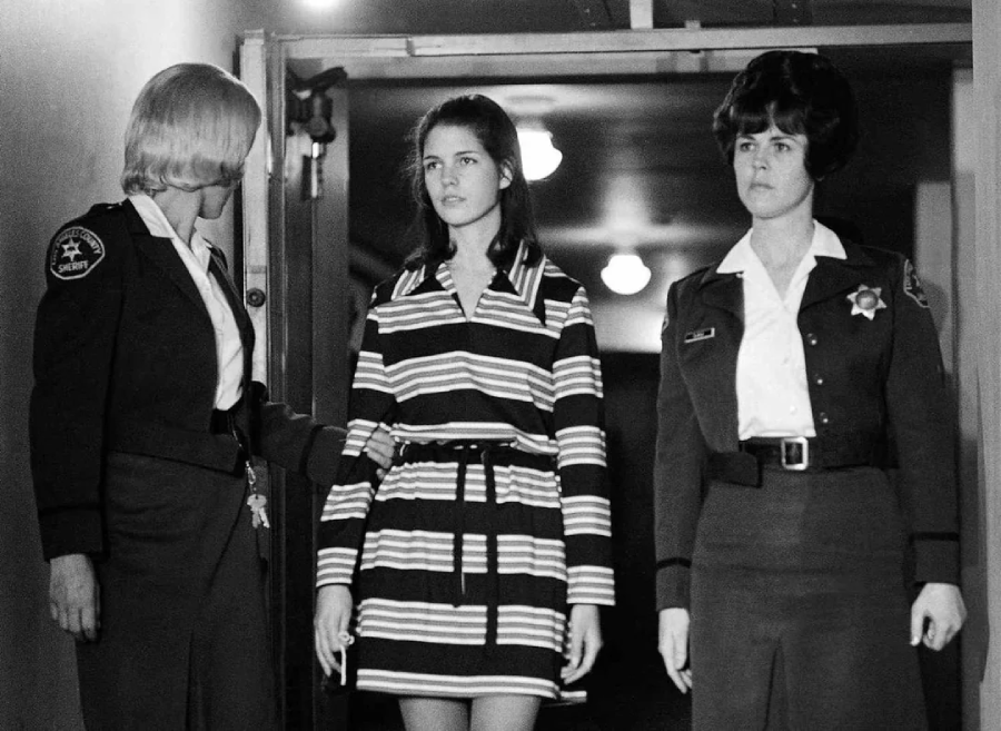 A young woman in a striped dress walks between two uniformed female officers in a hallway, with lights visible on the ceiling behind them. The officers each hold one of her arms.