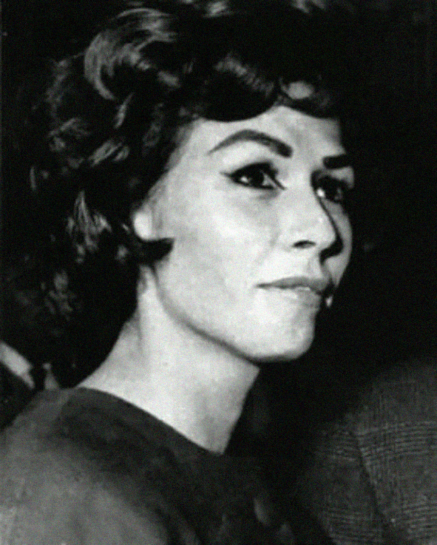 Black and white photo of a woman with short, wavy dark hair and bold eyeliner, looking slightly to the side with a neutral expression. She is dressed formally and appears to be at an event.