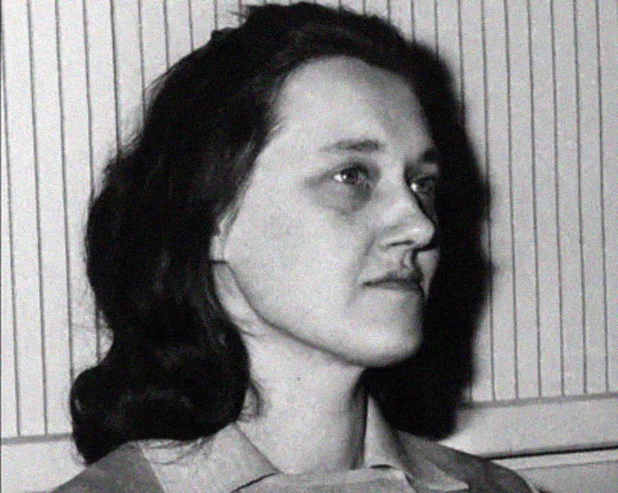 Black and white photo of a woman with dark, wavy hair, looking slightly to the right. She has a neutral expression and is wearing a collared garment, with a striped wall in the background.