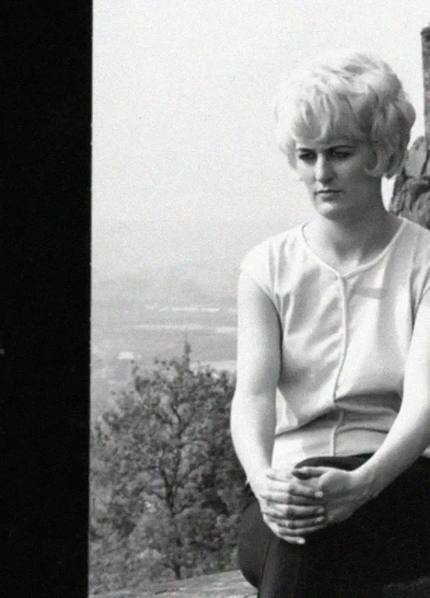 A woman with short, light-colored hair sits outdoors, looking down with a serious expression. She wears a sleeveless top and has her hands clasped on her knee. Trees and a hazy landscape are visible in the background.