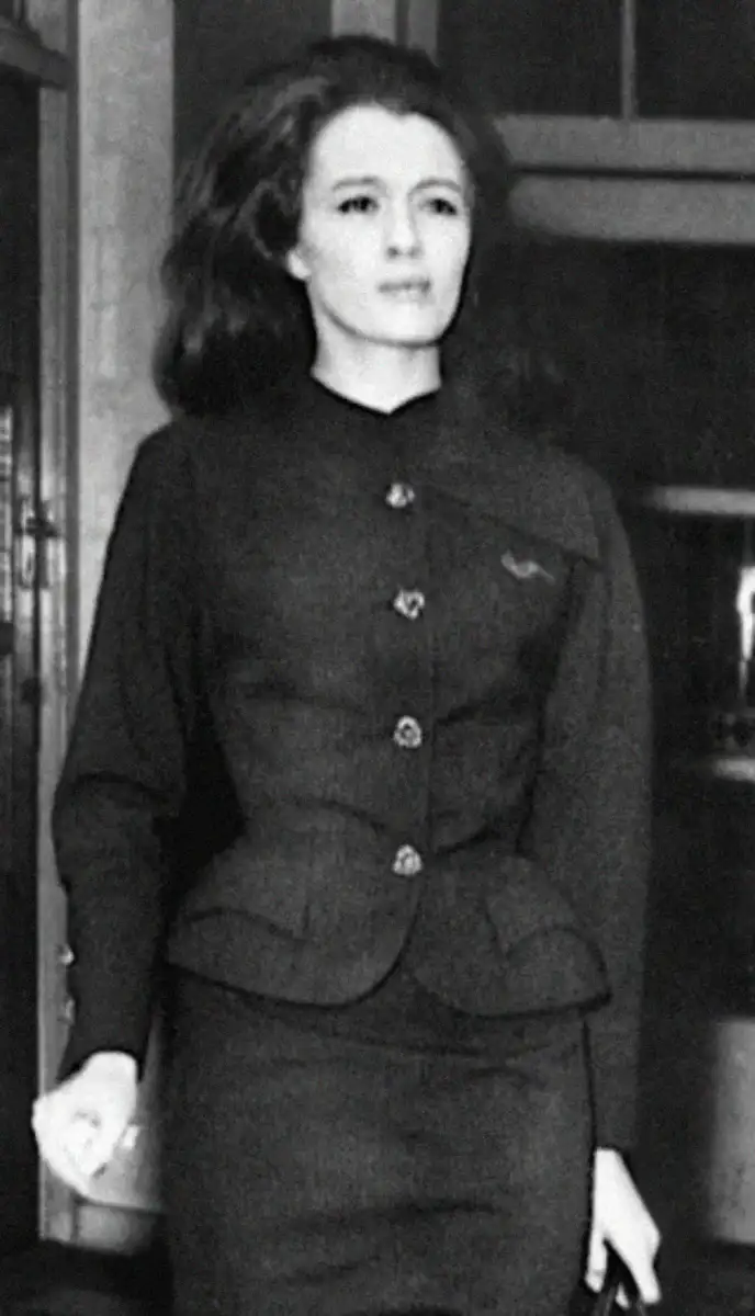 Black-and-white photo of a woman with long hair, wearing a dark, buttoned suit jacket and skirt, standing indoors, looking slightly to the side with a neutral expression.