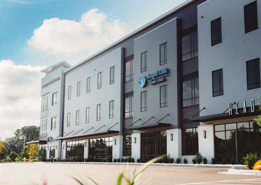 A modern three-story office building with several business signs, including Angel Oak Home Loans. The building has large windows, a parking lot in front, and some greenery along the sidewalk under a partly cloudy sky.