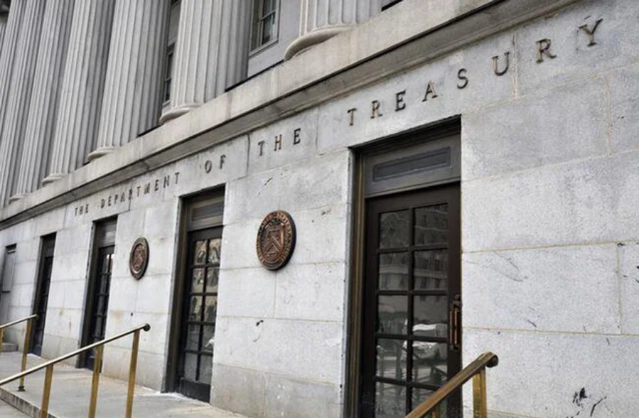 The exterior of the U.S. Department of the Treasury building, showing tall columns, stone walls, two entrances with metal doors, and round seals mounted next to the doors.