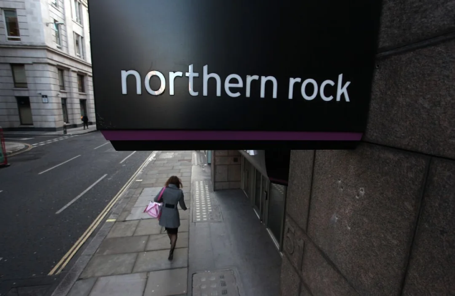 A person walks on a city sidewalk beneath a large sign reading "northern rock" attached to a building. The street is mostly empty and lined with stone buildings.