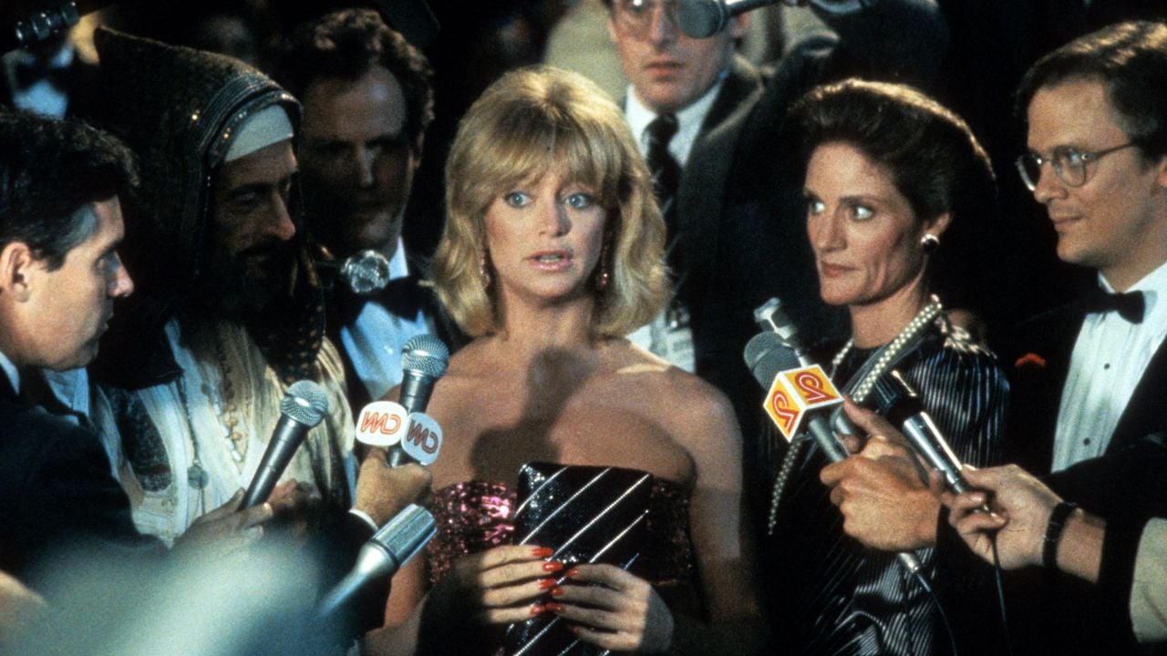 A blonde woman in a sparkly dress is surrounded by reporters holding microphones, with several people and cameras around her. She looks surprised or concerned as she stands in the middle of the crowd.
