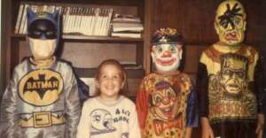 Four children stand side by side in front of a bookshelf, dressed in vintage Halloween costumes: Batman, a child in a white shirt, a clown, and Frankenstein&rsquo;s monster. All are smiling or posing for the camera.