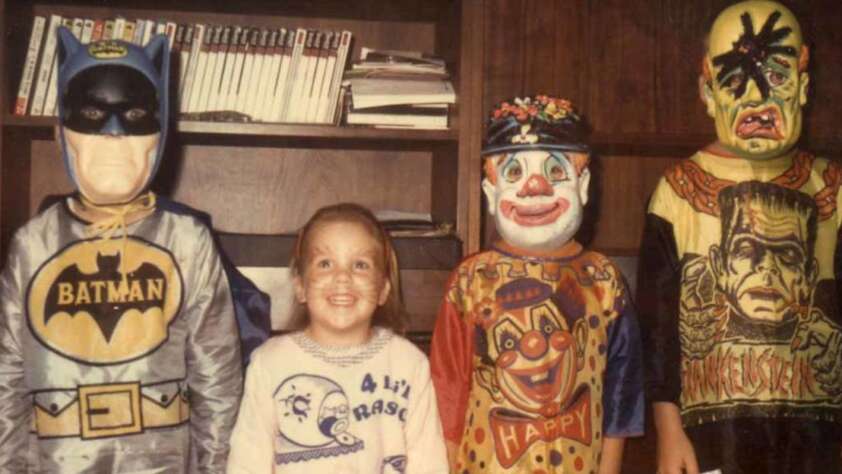 Four children stand side by side in front of a bookshelf, dressed in vintage Halloween costumes: Batman, a child in a white shirt, a clown, and Frankenstein’s monster. All are smiling or posing for the camera.