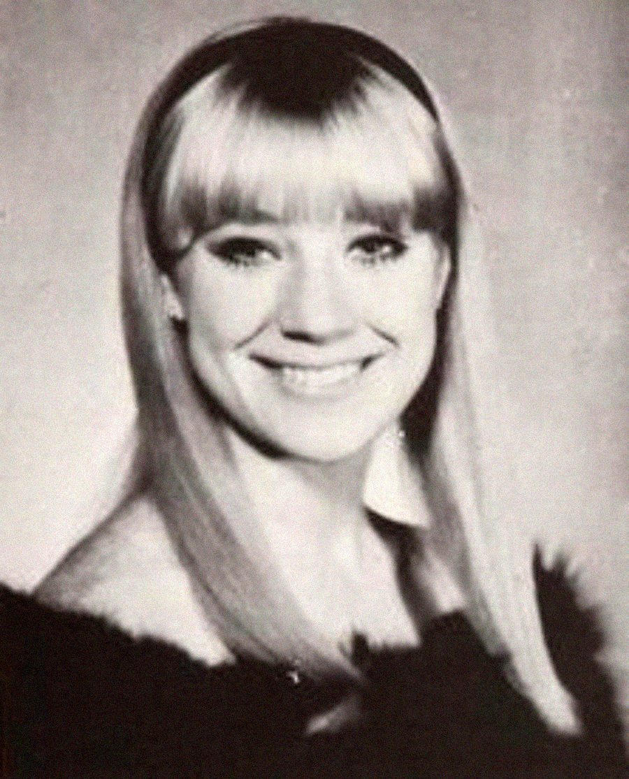 Black-and-white portrait of a smiling woman with long, straight, light-colored hair and bangs, looking slightly off to the side, wearing a dark off-the-shoulder top.
