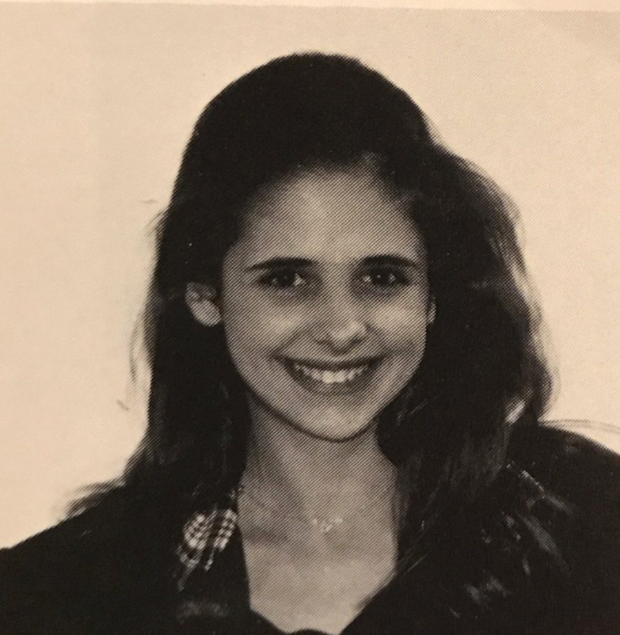 A young woman with long dark hair smiles at the camera. The image is black and white, and she is wearing a checkered-collar shirt partially visible under a dark jacket. The background is plain.
