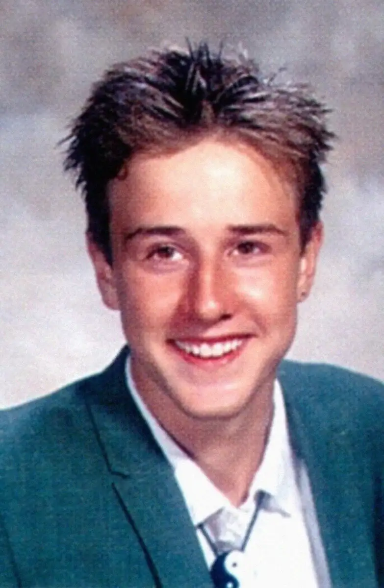 A teenage boy with short, spiky brown hair smiles at the camera, wearing a green blazer over a white collared shirt. The background is a soft, neutral color, typical of a school portrait.