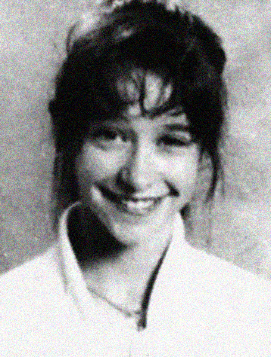 A black and white, slightly blurry portrait of a young woman with dark hair and bangs, smiling at the camera. She is wearing a light-colored top and a necklace. The background appears plain.