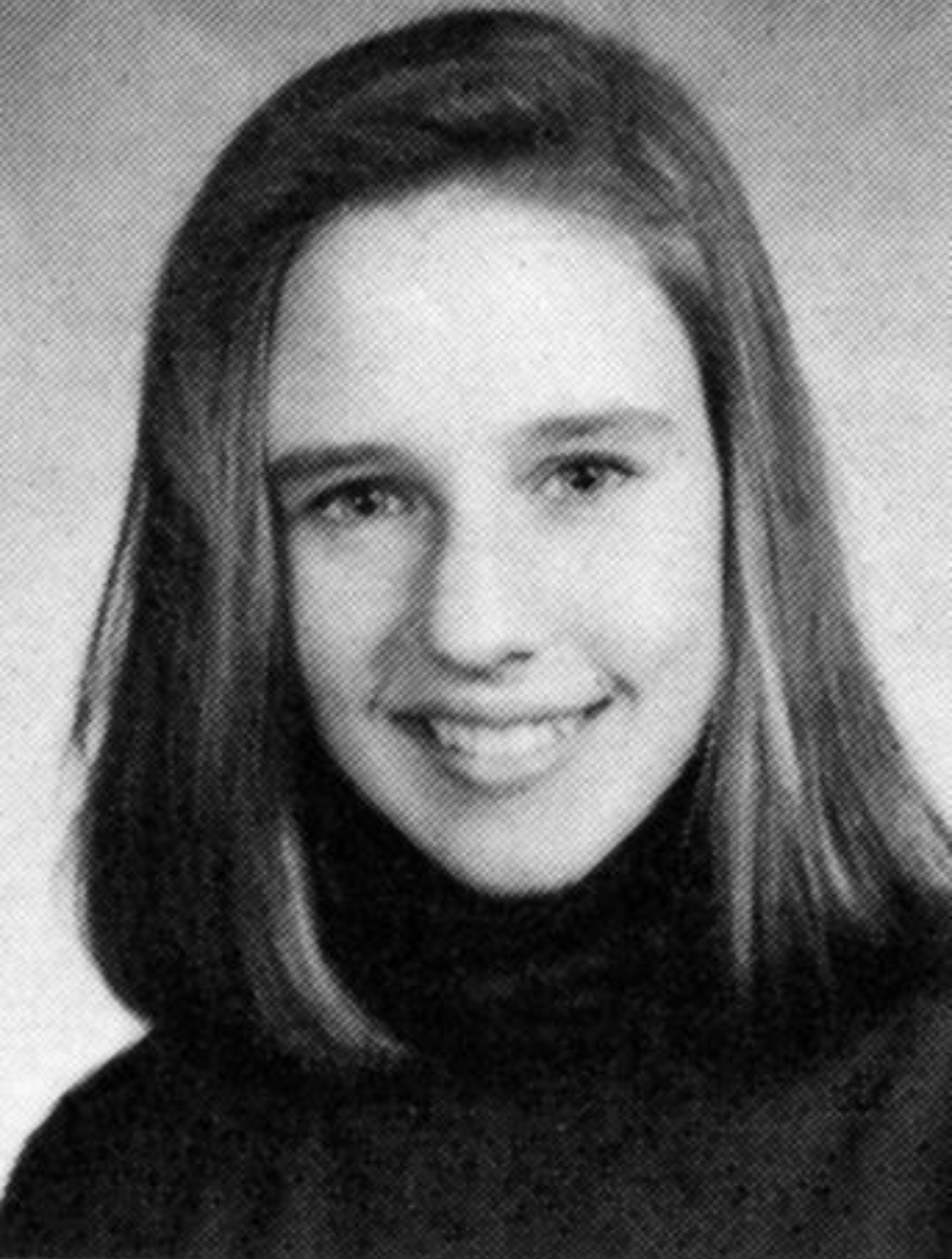 Black and white portrait of a young person with straight, shoulder-length hair, smiling and wearing a dark turtleneck sweater, posed in front of a plain background.