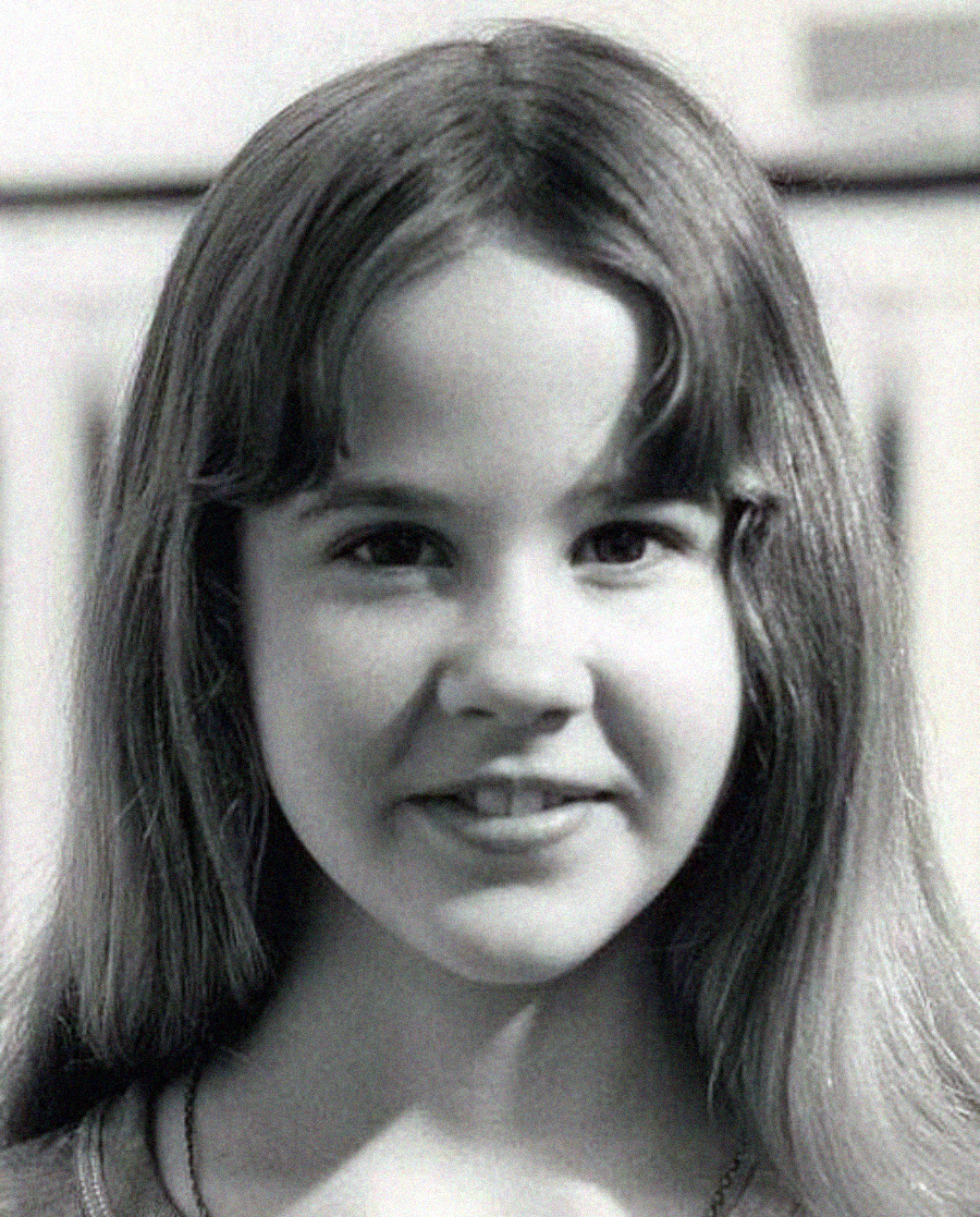 A black-and-white close-up photo of a young girl with long, straight hair and bangs, smiling softly and looking directly at the camera.