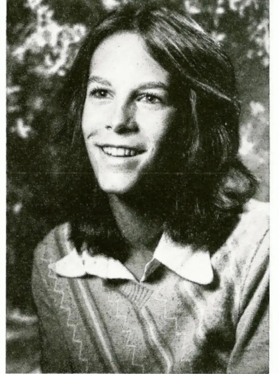 Black-and-white portrait of a young person with shoulder-length hair, wearing a light-colored collared shirt under a sweater, smiling and looking slightly to the side.