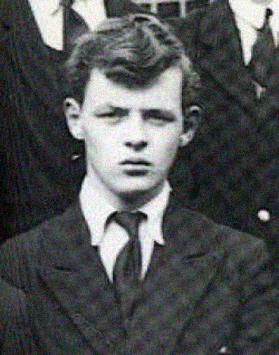 A black and white photo of a young man wearing a suit, tie, and collared shirt. He has short, wavy hair and is looking directly at the camera with a serious expression. Other people in suits are partially visible behind him.