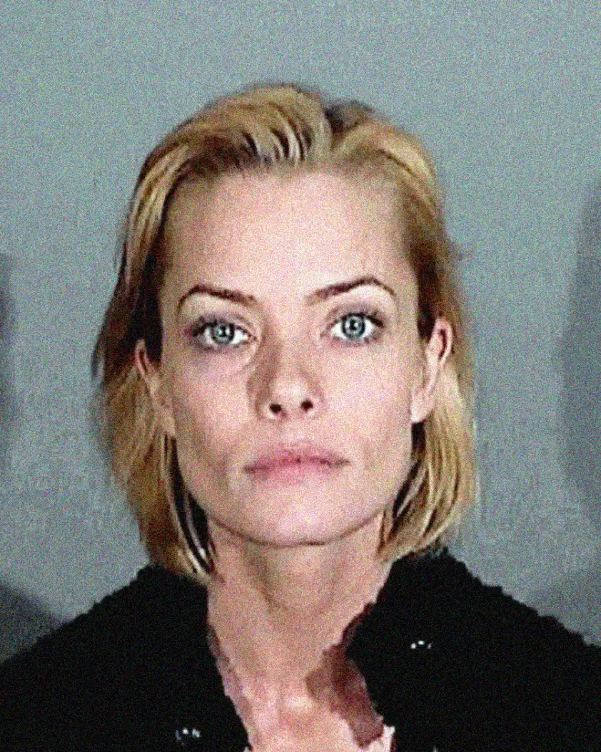 A woman with short blonde hair and light eyes stares directly at the camera with a neutral expression, against a plain, gray background. She is wearing a dark top.