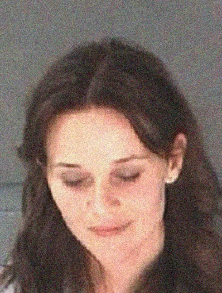 A woman with long, dark hair is looking down with a neutral expression. The image is slightly blurry and grainy, with a plain background.