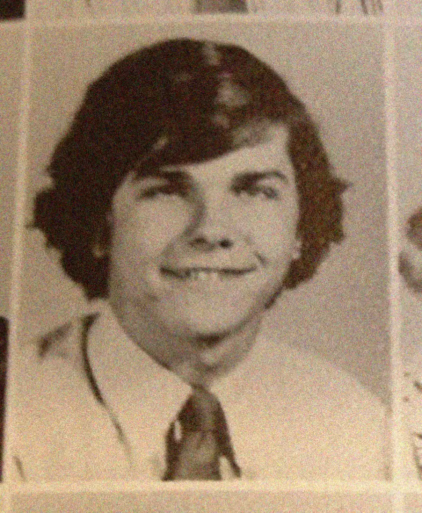 A black and white yearbook photo of a young man with medium-length dark hair, wearing a light-colored jacket, white shirt, and a tie, smiling slightly at the camera.
