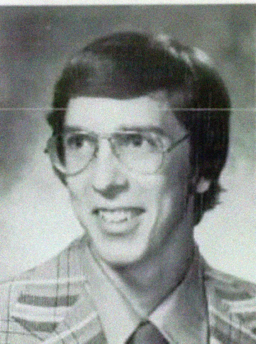 A black-and-white portrait of a young man with glasses, short dark hair, and wearing a plaid jacket, shirt, and tie, smiling at the camera.