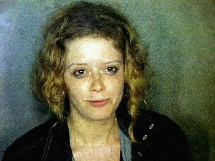 A woman with curly, shoulder-length blonde hair and pale skin looks at the camera with a neutral expression, wearing a dark jacket over a light top, against a plain greenish background.