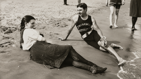 A woman and a man in vintage swimwear sit on wet sand at the beach, talking and smiling, while other people walk in the background near the water’s edge.