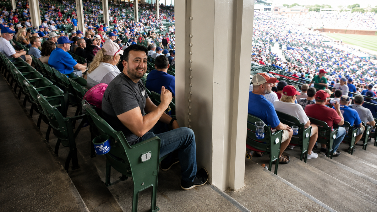 A man sits and smiles at a baseball stadium, giving a thumbs up. Fans fill the seats around him, while the field and more spectators are visible in the background. A pillar partially blocks the view beside him.