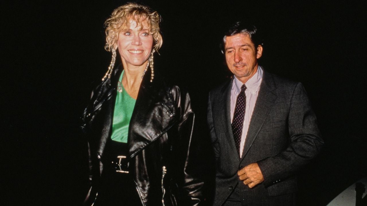A woman in a black leather jacket and green top smiles while walking beside a man in a suit and tie, both against a dark background.