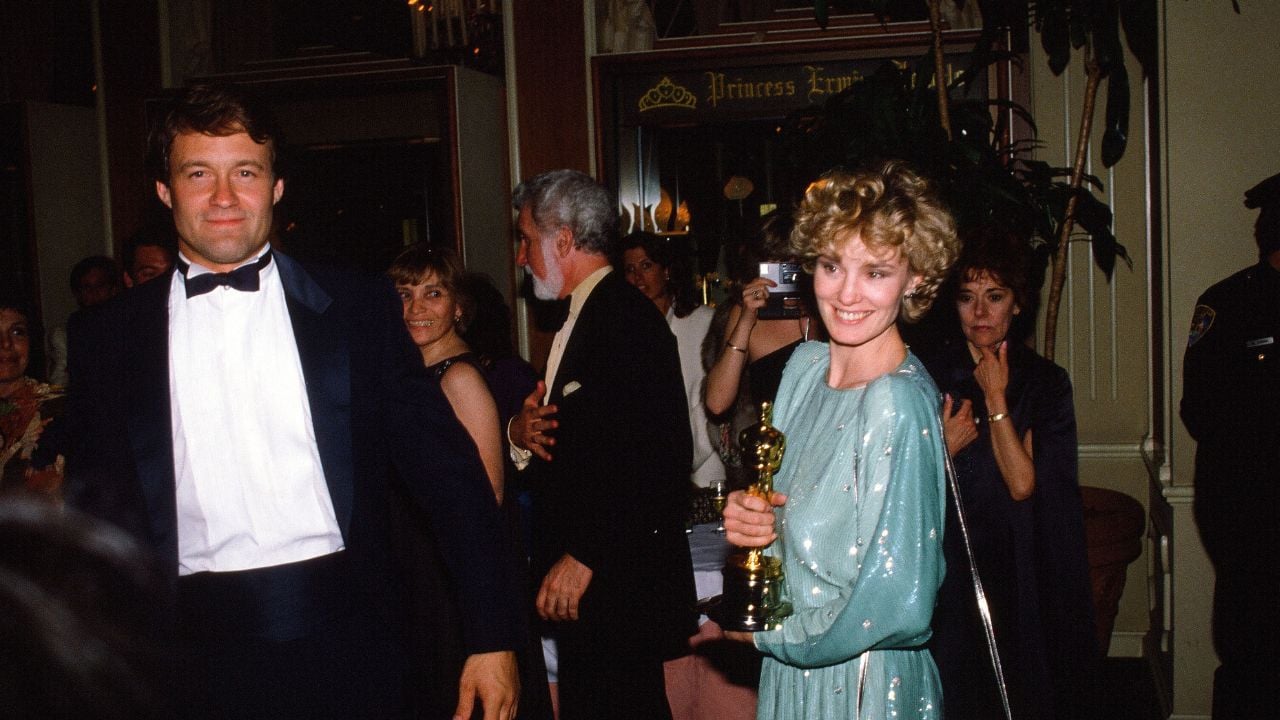 A woman in a light blue gown holds an Oscar trophy and smiles, standing next to a man in a tuxedo at a crowded, elegant indoor event. Other people in formal attire are visible in the background.