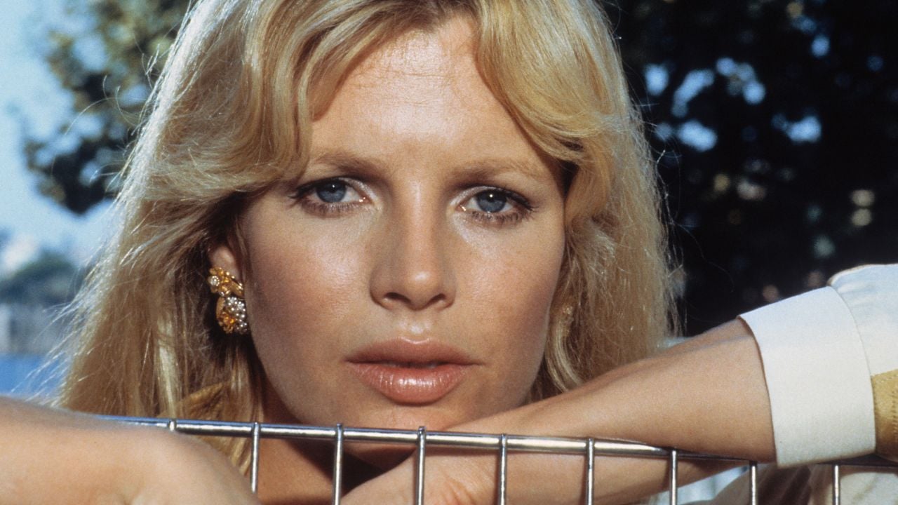 A woman with long blonde hair rests her arms on a metal fence, gazing directly at the camera. She wears gold earrings and a light-colored top, with a blurred outdoor background of trees.