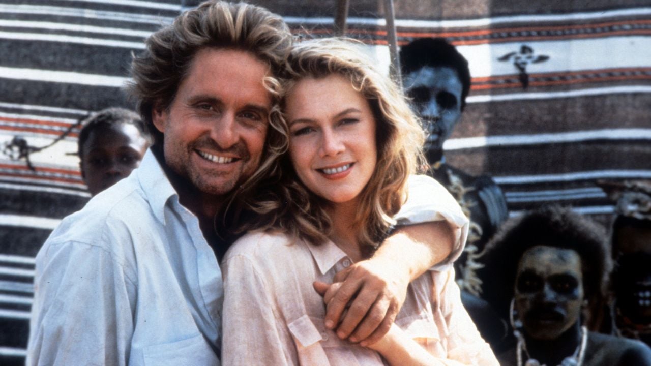 A man with wavy hair embraces a smiling woman in front of a striped textile, with several people in the background, some with faces painted white and dark.