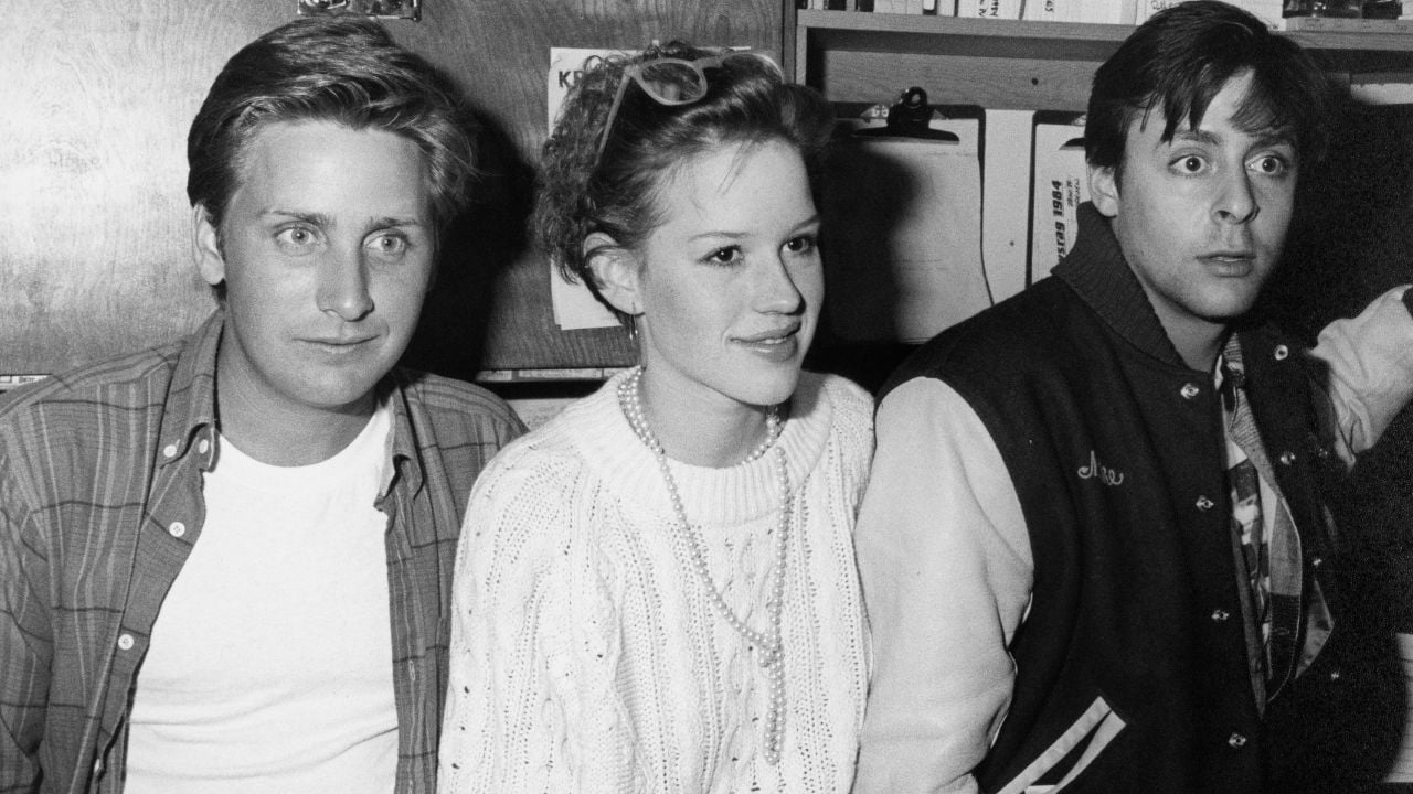 Three young adults sit close together. The person on the left wears a plaid shirt, the center person has glasses and a sweater, and the right person wears a varsity jacket and looks surprised. All are smiling or neutral.