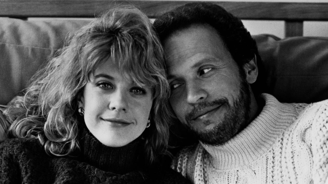 A woman with wavy hair smiles at the camera while a bearded man in a sweater leans next to her, looking at her affectionately. Both are sitting close together on a sofa.