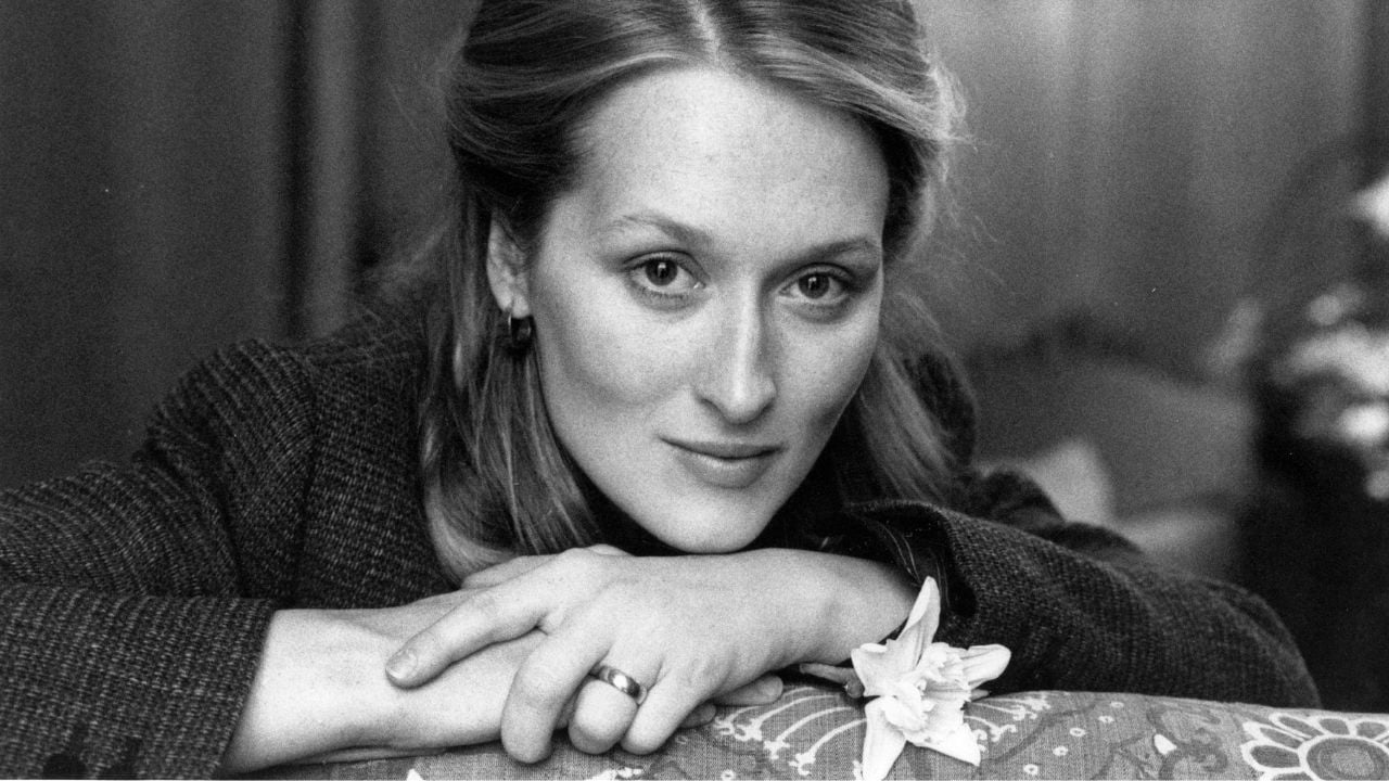 A woman with light hair rests her chin on her hands, looking at the camera. She wears a ring and a dark top, with a flower near her hand. The image is in black and white.