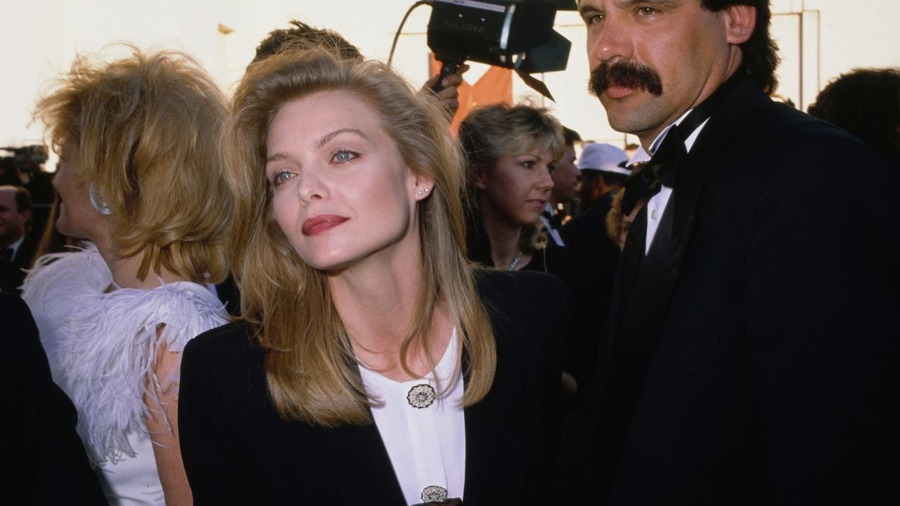 A woman with long blonde hair in formal attire stands among a crowd at an event, looking slightly to the side. Other well-dressed people, some with cameras, appear in the background.