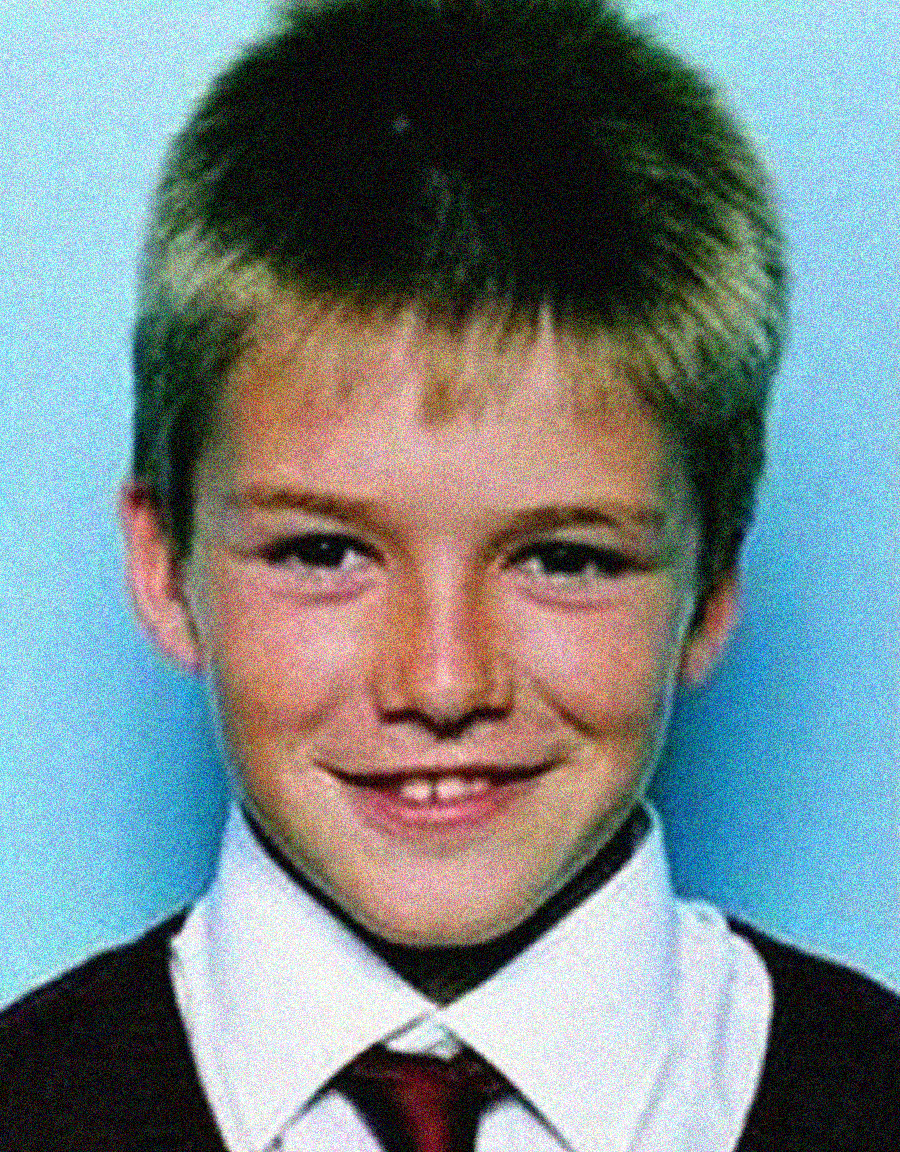 A young boy with short, light brown hair smiles at the camera. He is wearing a white shirt, a red tie, and a dark sweater. The background is a solid light blue.