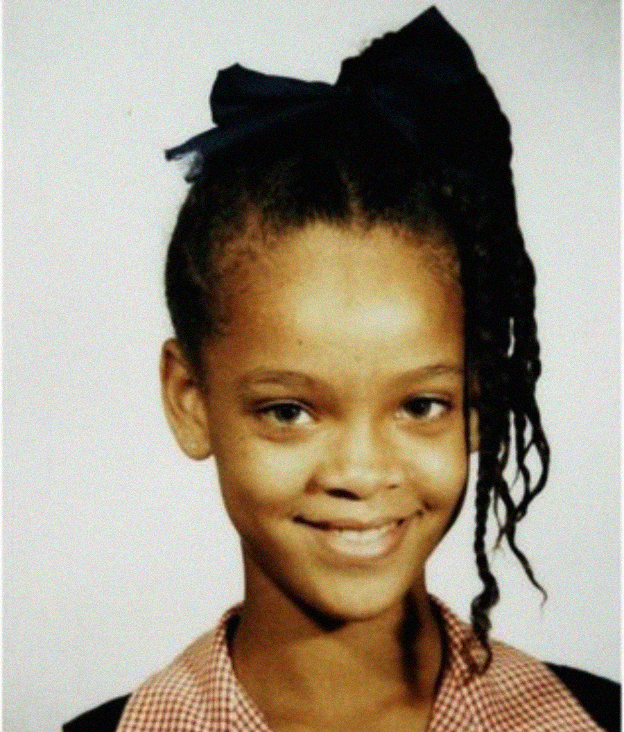 A young girl with a big navy blue bow in her hair smiles at the camera. She is wearing a red-checked uniform dress with a cream collar, and her hair is styled in braids. The background is plain and light-colored.