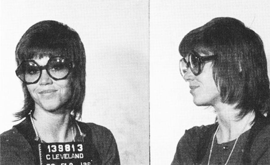 A black-and-white mugshot photo showing a woman with large round glasses and shoulder-length hair, facing forward and in profile. She is wearing a dark top and a necklace, with a booking board labeled "CLEVELAND.
