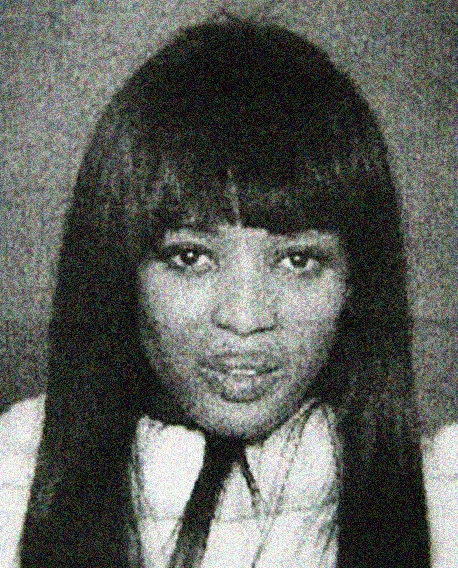 A black and white portrait of a woman with long, straight hair and bangs, looking directly at the camera with a neutral expression. She is wearing a light-colored top.