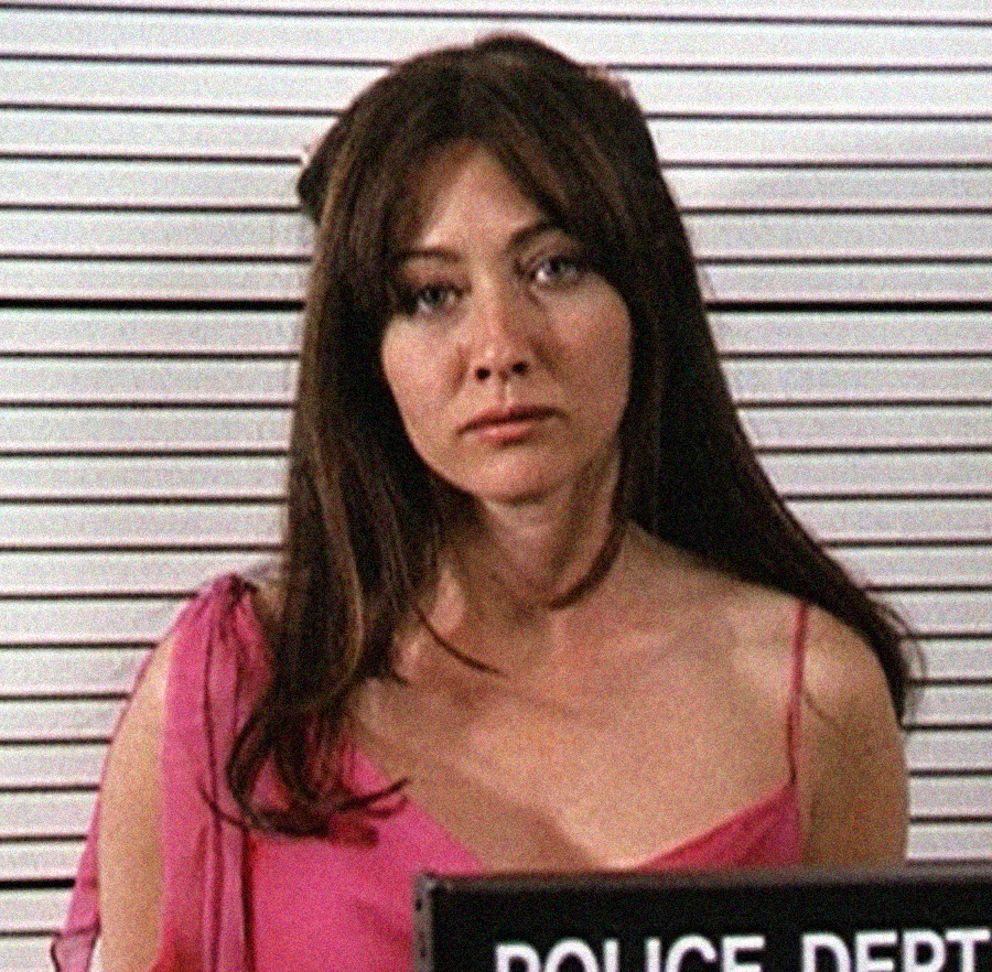 A woman with long brown hair wearing a pink top stands in front of a height chart holding a police department sign, appearing to be in a mugshot.