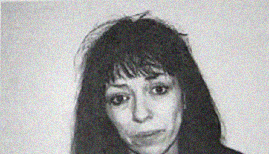 A black-and-white photo of a woman with long dark hair and bangs, looking directly at the camera with a neutral expression. The background is plain and light-colored.