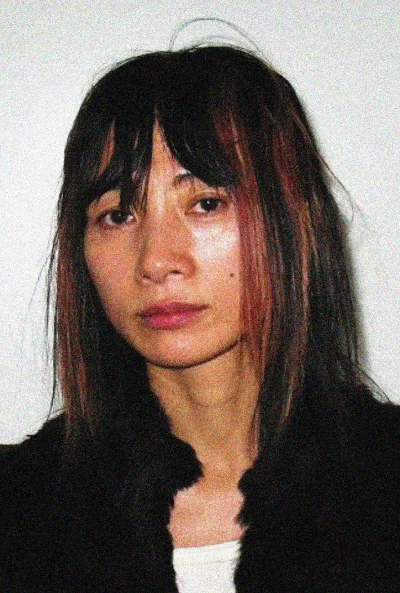 A person with straight, shoulder-length dark hair and reddish highlights looks at the camera with a neutral expression, standing against a plain white background.
